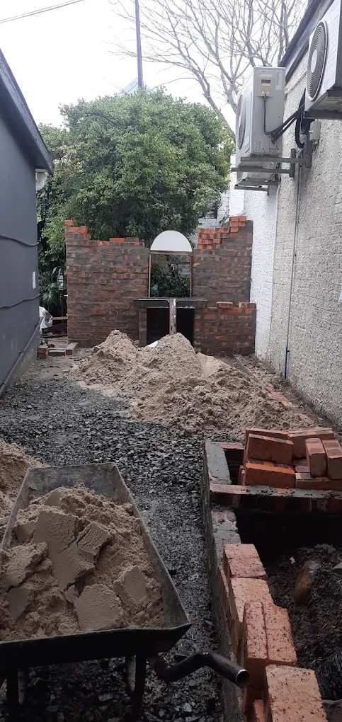 A clay-brick braai under construction between two houses, with the arched opening formed and sand, gravel and stacked bricks on site.