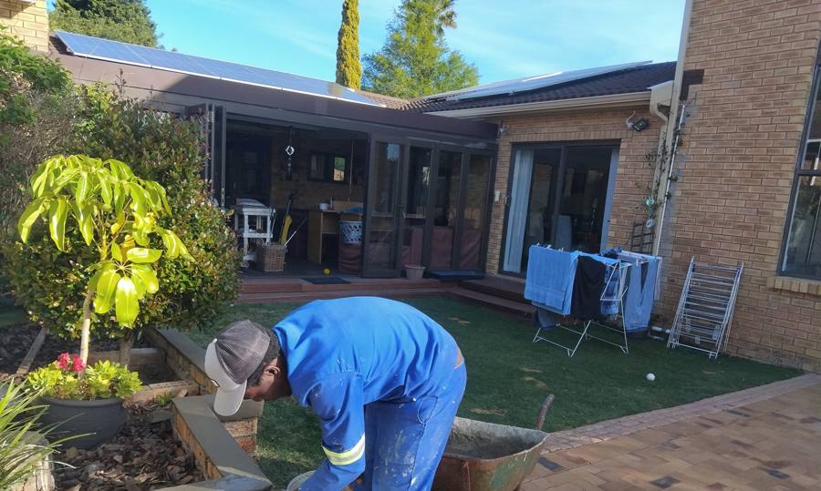 A bricklayer bedding a brick in mortar on a low garden wall, with a wheelbarrow beside him and a glazed covered patio across the lawn behind.