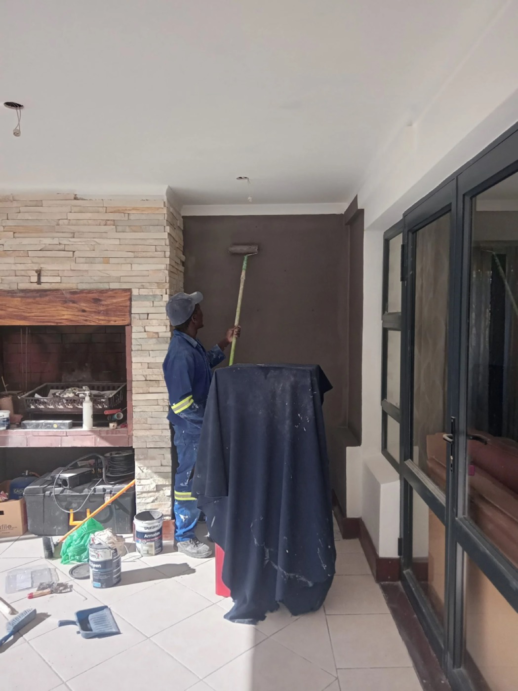 A painter in overalls rolling a fresh coat of paint onto an interior wall, with drop sheets and paint tins on the tiled floor